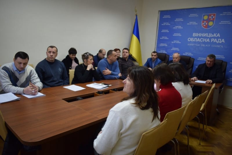 Група людей сидить за столом перед прапором.