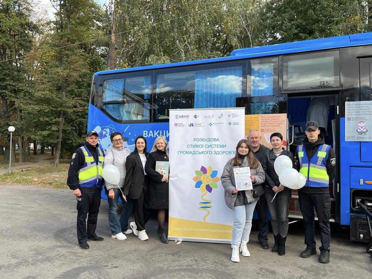 Групове фото п'ять жінок і три чоловіки біля банера з текстом"Розбудова стійкої системи громадського здоров'я"