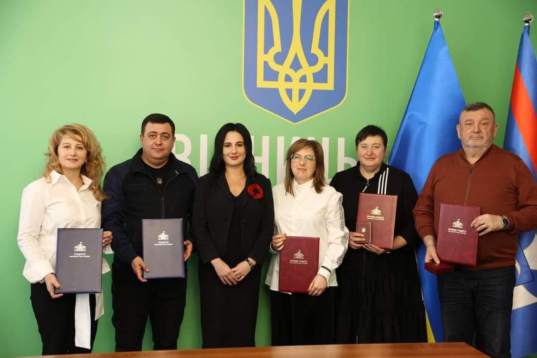 Люди стоять перед зеленим фоном, тримаючи у руках нагороди.