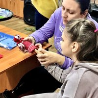 Жінка і дівчинка сидять поруч за столом, увага обох зосереджена на ляльці-мотанці, яку вони тримають у руках.