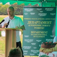 Директор Департаменту агропроммислового розвитку стоїть за трибуною у тимчасовому павільйоні посеред поля трмамає мікрофо 