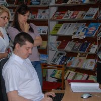 Чоловік сидить перед комп'ютером. Дві жинки столять поруч і дивляться на екран комп'ютера