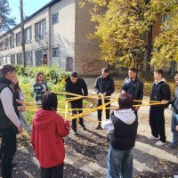 Група молодих людей стоять на вулиці, утворивши коло. Усі тримають у руках яскраво-жовту мотузку, яка переплетена таким чином, що утворює складну фігуру, схожу на багатокутну зірку.