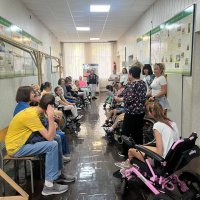 Діти і дорослі сидять у довгій кімнаті і слухають доповідача.Серед учасників присутні діти на інвалідних візках.